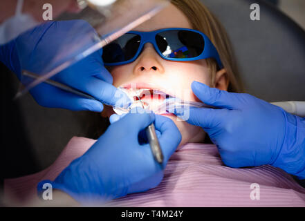 Dentist performing dental filling procedure to a little girl in pediatric dental clinic with a help of assistant. Calm child is sitting in a dental ch Stock Photo