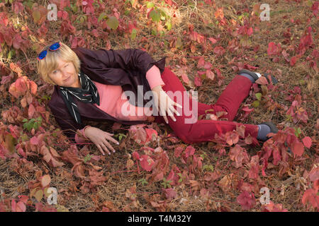 Happy middle-aged woman lying on the grass in early autumn. Pink clothes. There are large pink leaves around. Stock Photo