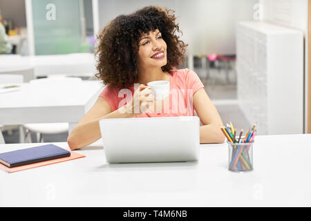 Young cheerful business woman on laptop computer in the office drinking a cup of coffee Stock Photo