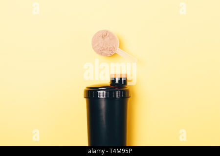 Protein powder in measuring spoon and black shaker on yellow background, top view. Stock Photo