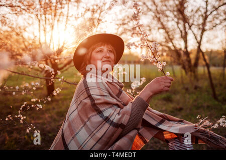 Happy middle-aged woman relaxing in spring blooming garden at sunset. Woman walking and dancing with gift box. Mother's day concept Stock Photo