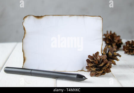 On white wooden boards, a sheet of paper burned at the edges, forest cones, a black pen at the edges. Leaving space for text. Concept background. Stock Photo