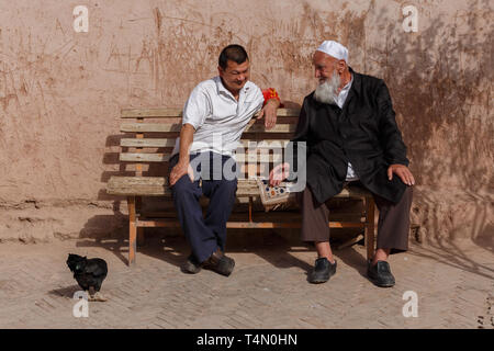 Two Uyghur men having a conversation on a bench. Notable detail: the chicken on the left side of the two gentlemen. Kashgar, Xinjiang Province, China. Stock Photo