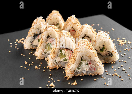 Tasty triangle california uramaki filled with cream cheese, cucumber and artificial crab meat. Sushi covered with sesame, presented on black stone slate plate. Stock Photo