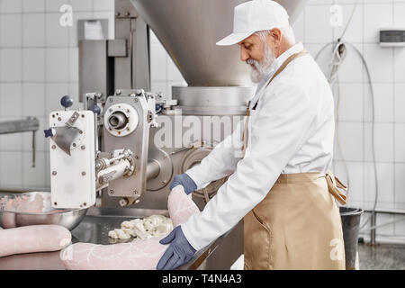 Butcher man working with production of sausages, standing near equipment, holding sausage. Worker wearing in white uniform, brown apron, rubber gloves. Food industry. Stock Photo