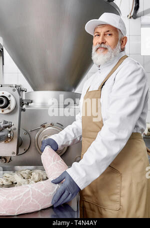 Elderly professional worker in white uniform and gloves doing sausages using special equipment. Butcher man holding big sausage, looking at camera, posing. Meat production. Stock Photo