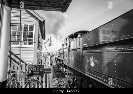 Black & white scene of railway signalman at vintage station looking out of signal box window greeting cab crew of puffing vintage steam train passing. Stock Photo