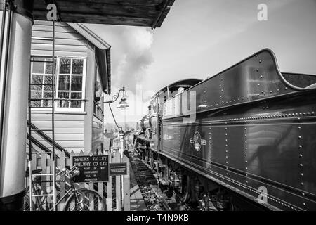 Black & white railway scene, signalman at vintage station looking out of signal box window waving to cab crew on puffing vintage steam train passing. Stock Photo