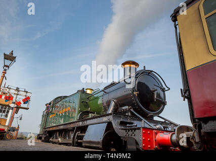 Low angle, front view close up of old vintage UK steam locomotive in morning spring sunshine puffing as it waits alongside platform at railway station. Stock Photo