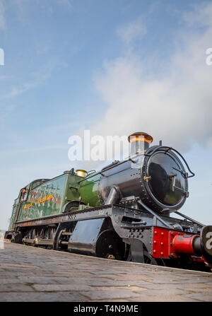 Low angle, front view of vintage UK steam locomotive in morning sunshine waiting by platform at railway station. Stock Photo