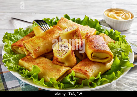 close-up of Savory crepe rolls with ground chicken meat and champignon filling served on a bad of fresh lettuce leaves on a white plate on a wooden ta Stock Photo