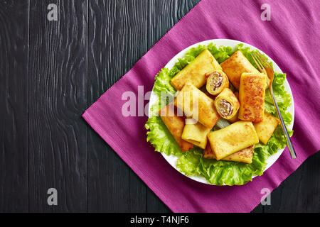 overhead view of Savory crepe rolls with ground chicken meat and champignon filling served on a bad of fresh lettuce leaves on a white plate on a blac Stock Photo