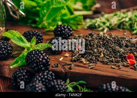 Heap of Dry Green Tea and Fresh Blackberries on Wooden Cutting Board. Bundles of Mint and Thyme Leaves. Stock Photo