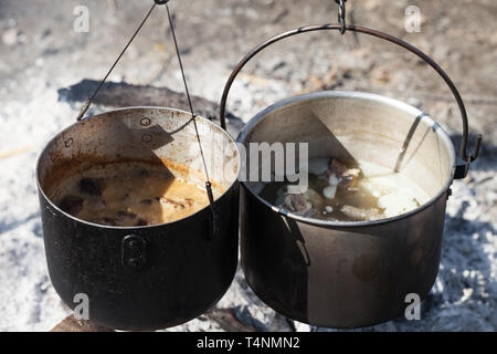 Cooking in two sooty old cauldrons on campfire at sunny day Stock Photo