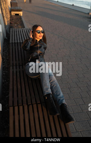 Young woman listens to music in closed headphones through her phone wearing a leather jacket and jeans at a sunset near river Daugava Stock Photo