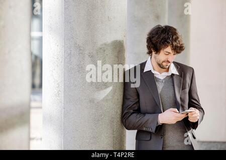 Young man leaning on column, looks on smartphone in hand, North Rhine-Westphalia, Germany Stock Photo