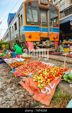 Samutsakorn Province, Thailand - March 14, 2019: Daily life of a street vendor selling vegetables between train tracks as train dangerously passes by. Stock Photo