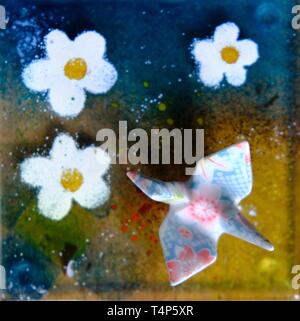 Close up overhead view of decorative glass art featuring fused white flowers and a colorful patterned origami bird over a blue and green background Stock Photo