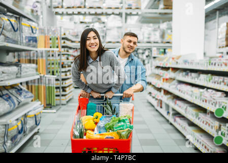 Couple with cart full of goods in a supermarket, family shopping. Customers in shop, buyers in market Stock Photo