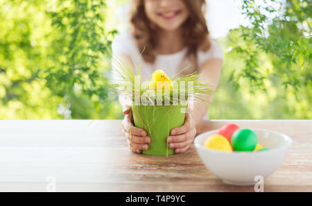 easter, holiday and child concept - close up of girl with toy chicken in flowerpot with grass and colored eggs over green natural background Stock Photo