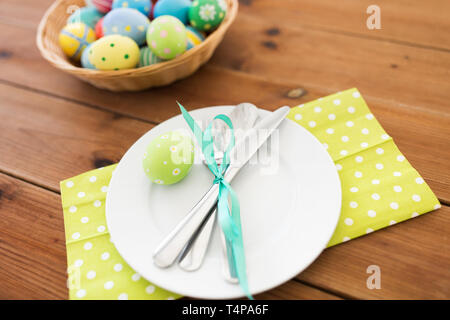 easter, holidays and table setting concept - close up of plate with cutlery and colored eggs on wooden table Stock Photo