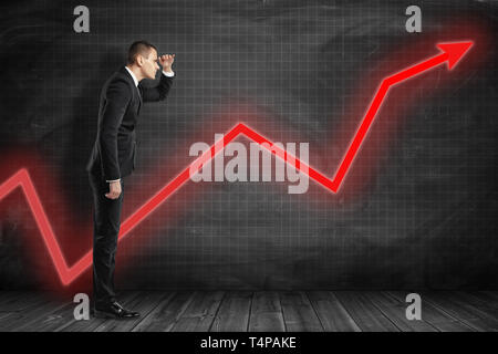 Side view of businessman with hand at forehead peering at something in the distance standing near black wall with red arrow going up on it. Stock Photo