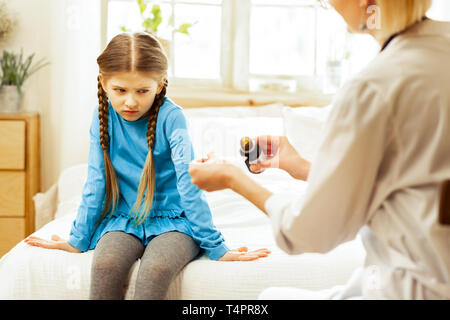 Unhappy about medication. Small sick upset pretty girl with long brown braids dissatisfiedly looking at a pediatrician in a white coat pouring throat  Stock Photo