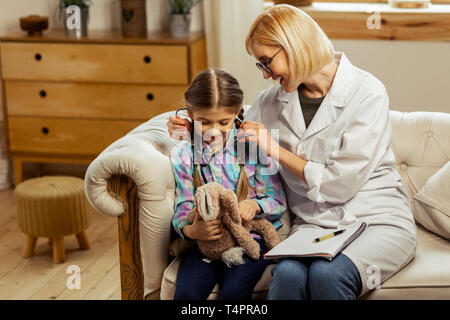 A sick child with a doctor. Charming beautiful smiling cheerful short-haired physician giving a sick cute girl a stethoscope to try on while sitting o Stock Photo