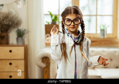 Pretending a doctor. Small cute sweet brown-haired girl with long braids trying on a costume of a doctor with glasses. Stock Photo