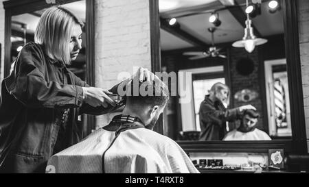 Young woman making haircut for bearded man at barbershop. Female barber at salon. Gender equality. Woman in the male profession. Black and white or colorless photo. Hairstyle, salon, hairdresser, lifestyle concept. Stock Photo