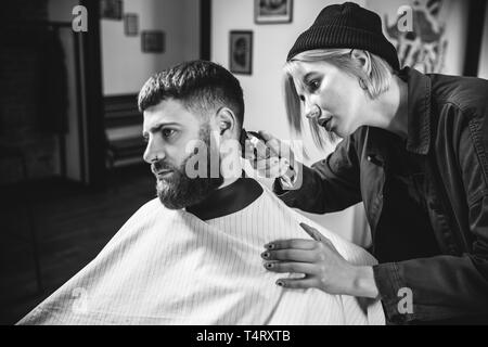 Young woman making haircut for bearded man at barbershop. Female barber at salon. Gender equality. Woman in the male profession. Black and white or colorless photo. Hairstyle, salon, hairdresser, lifestyle concept. Stock Photo