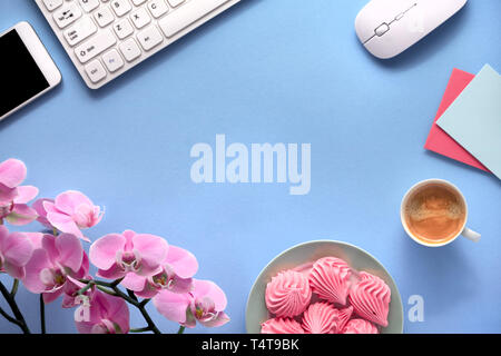 Pink phalenopsis orchid, greeting cards, keyboard, mobile phone, plate of marshmallow and cup of coffee. Flat lay on blue paper background with copy-s Stock Photo