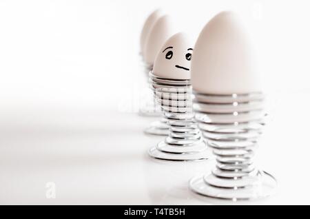 A line of four white chicken eggs in silver egg cups, one with thoughtful facial expression. Stock Photo
