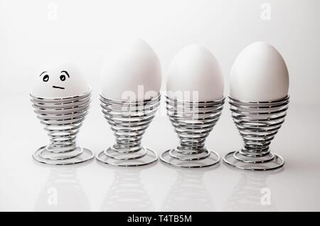 Four white chicken eggs in silver egg cups, one anthropomorph chicken egg with grumpy facial expression, in silver egg cups. Stock Photo
