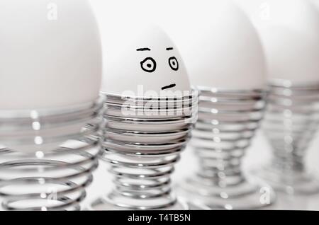 Four white chicken eggs in silver egg cups, one anthropomorph chicken egg with grumpy facial expression, in silver egg cups. Stock Photo