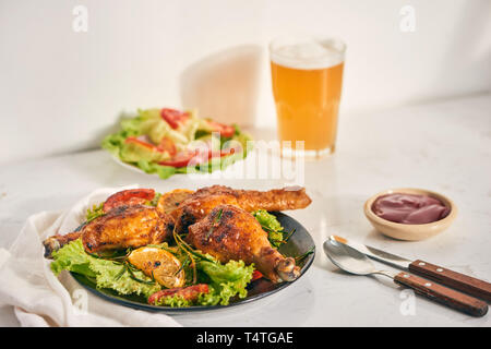 Grilled chicken legs roasted on the grill on dark plate with tomato sauce in a bowl and lettuce leaves, glass mug of beer Stock Photo