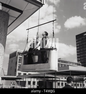 1960s, historical, two window cleaners standing in a concrete hoist with bucket, suspended from above, looking up at the tall office block they will have to clean by hand, London, England, UK. Stock Photo