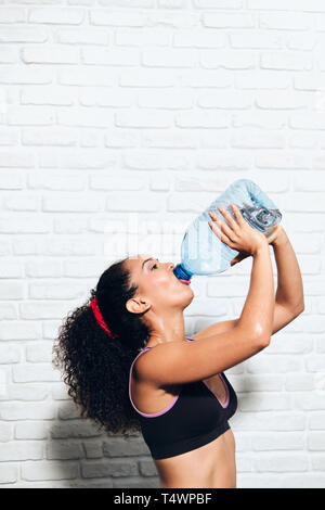 Portrait of tired african american woman drinking gallon of water. Thirsty black girl with big bottle in gym rehydrating after working out. Stock Photo