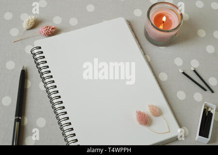 Top view of a notebook with a blank page, a pen, black wooden matches, coral candles, dried flowers on grey table with white dots. Stock Photo