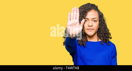 Young beautiful woman with curly hair wearing winter sweater doing stop sing with palm of the hand. Warning expression with negative and serious gestu Stock Photo
