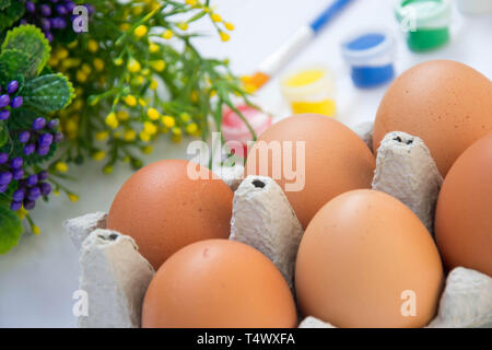 Easter brown chicken eggs in carton container, with plastic flowers and blurred paints, colorful palette on the background, paint equipment on white b Stock Photo