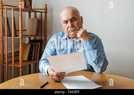 Thoughtful old man reading a handheld paper document with a look of concentration as he sits indoors at a table Stock Photo
