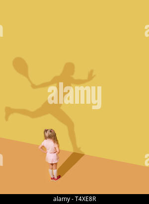 Faster and stronger than others. Childhood and dream concept. Conceptual image with child and shadow on the yellow studio wall. Little girl want to become tennis player, champion, winner, sportswoman. Stock Photo