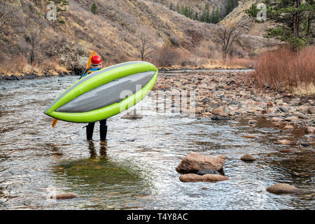 paddler carrying inflatable whitewater kayak on a shore of mountain river in early spring - Poudre River above Fort Collns, Colorado Stock Photo