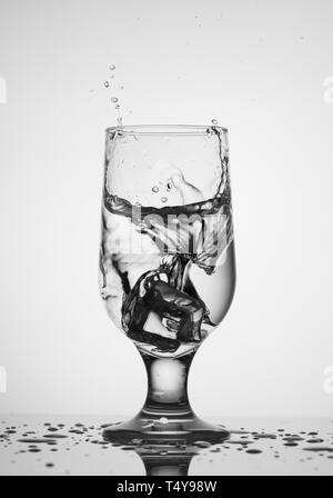 The cube of ice falls into a clear glass glass with water. Splash of small drops of water on the walls of the glass and on the table. Stock Photo