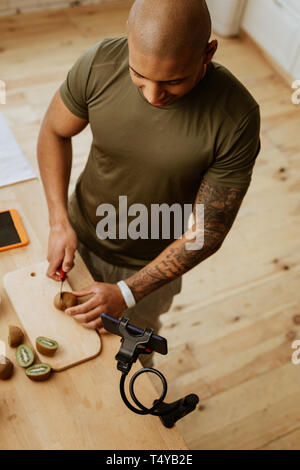 Sportsman with tattoo cutting kiwi while filming video about food Stock Photo