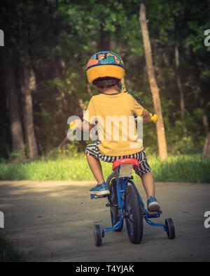 Vintage tone rear view of Asian toddler boy riding tricycle at nature park near Dallas, Texas, USA. Healthy kid cycling with helmet, short and sneaker Stock Photo