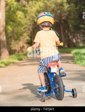 Rear view of Asian toddler boy riding tricycle at nature park near Dallas, Texas, USA. Healthy kid cycling with helmet, short and sneaker on park conc Stock Photo