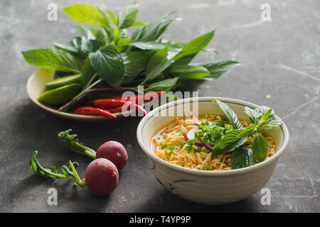 Instant noodles in bowl with fresh herbs, garnish of cilantro and Asian basil, lemon, lime on dark stone background Stock Photo