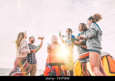 Group of friends having barbecue drinking beers while camping on the beach - Happy people enjoying camp bbq playing guitar and listening music Stock Photo
